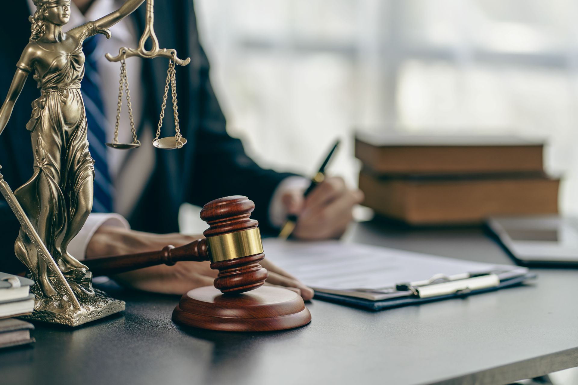 Gavel and scales of justice on desk beside person writing on legal documents. Gavel and scales of justice on desk beside person writing on legal documents.
