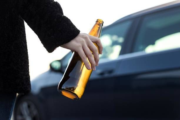 A person is holding a bottle of beer in front of a car