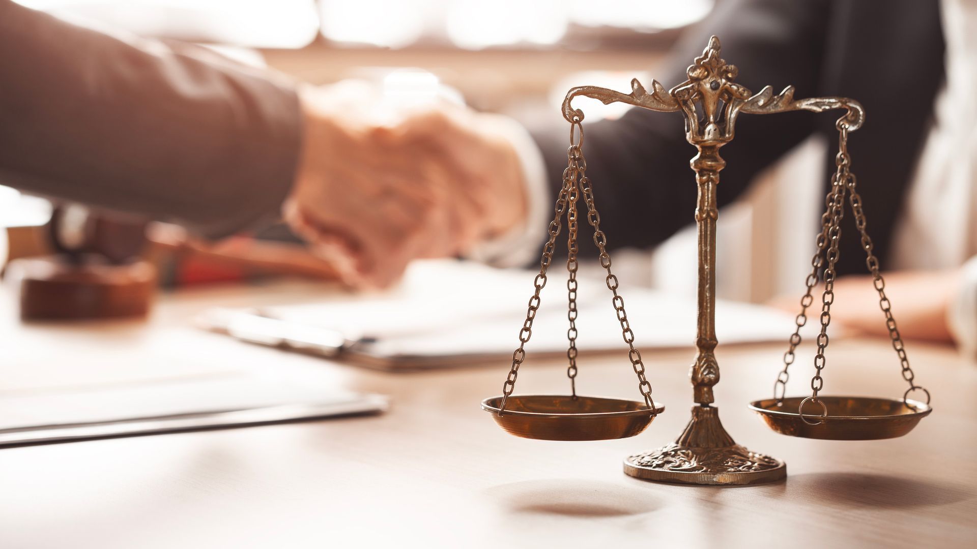 Close-up of justice scales on a desk with two people shaking hands in the background.