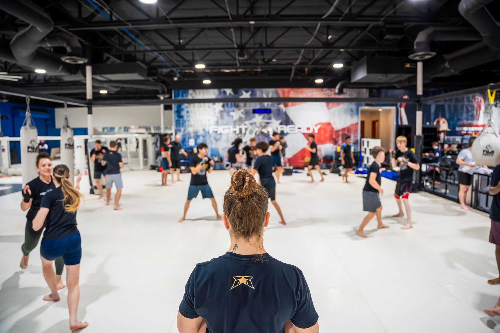 People practicing martial arts in a bright gym, seen from behind a coach.