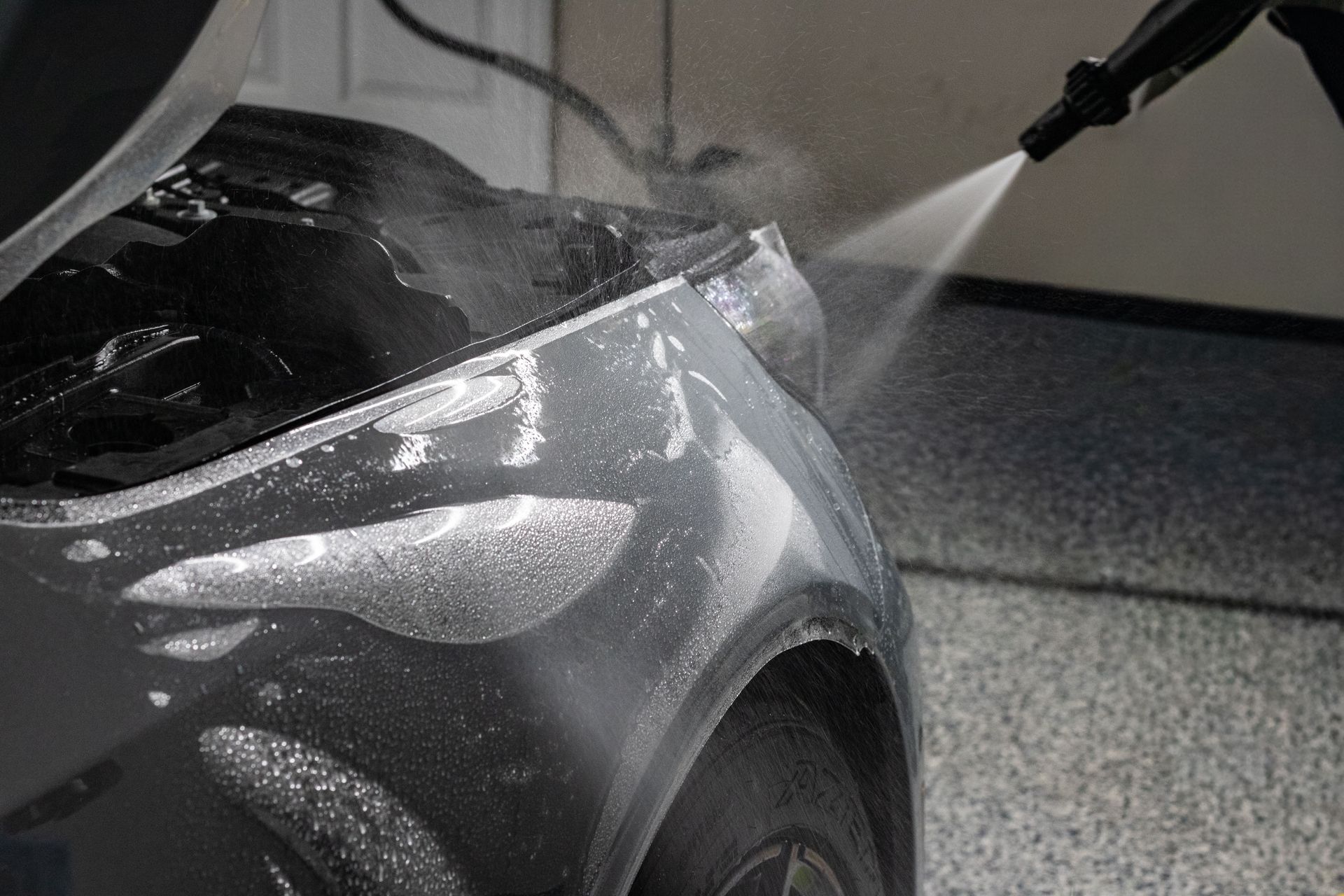 Close-up of a car being pressure washed, with water spraying over the hood and front wheel.