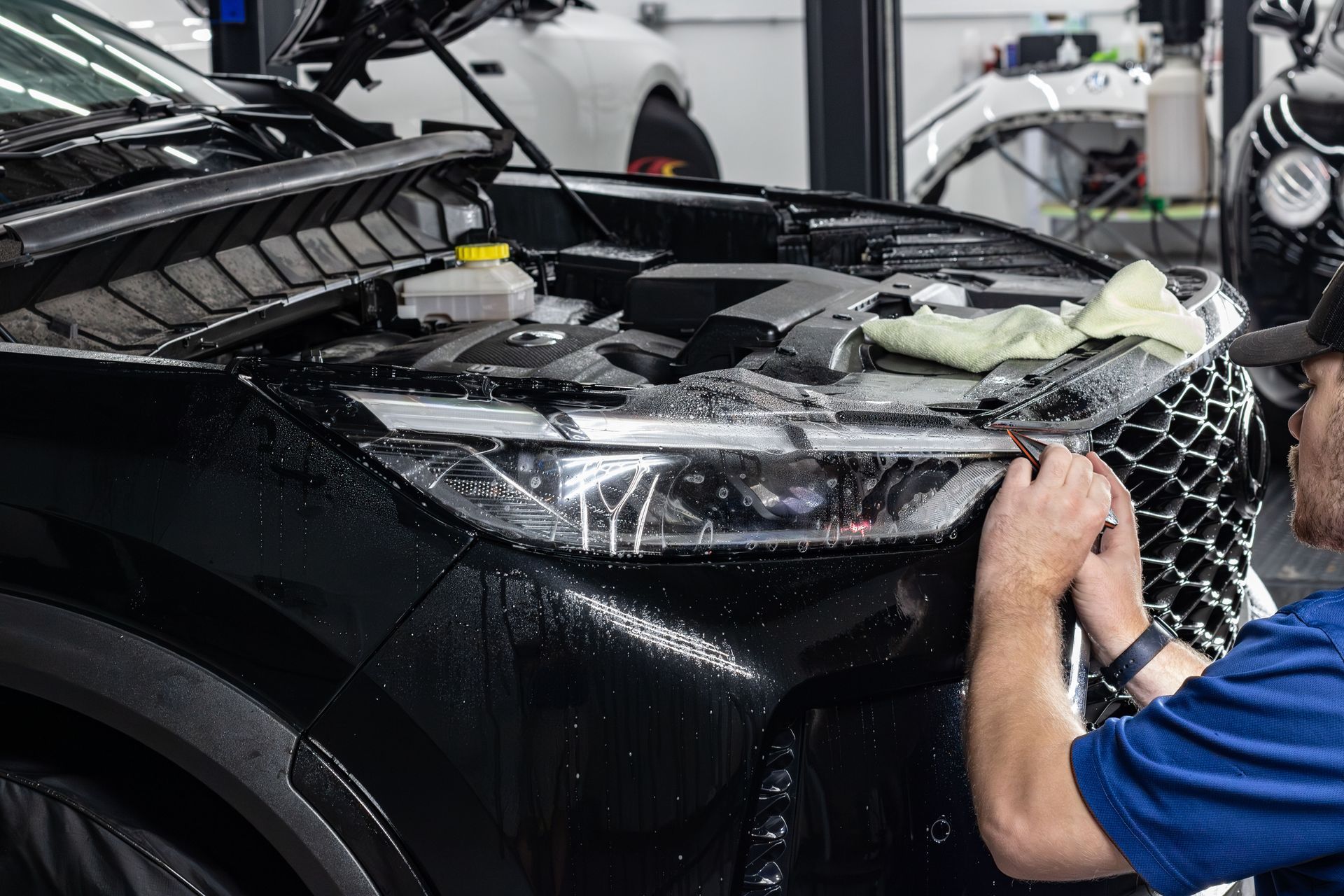 Mechanic repairing a black car’s front end in a garage, with the hood open and parts exposed