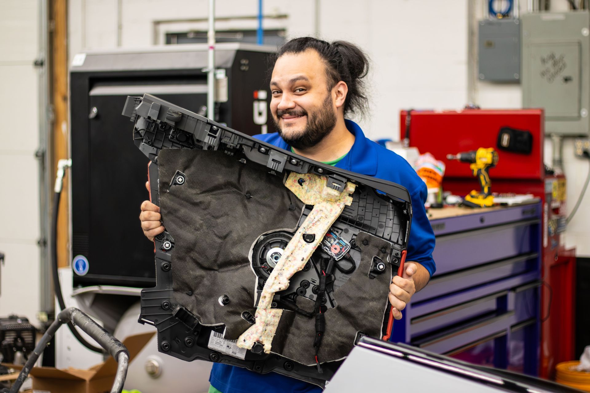 Smiling mechanic holding a large car door panel in a workshop, with tool cabinets in the background