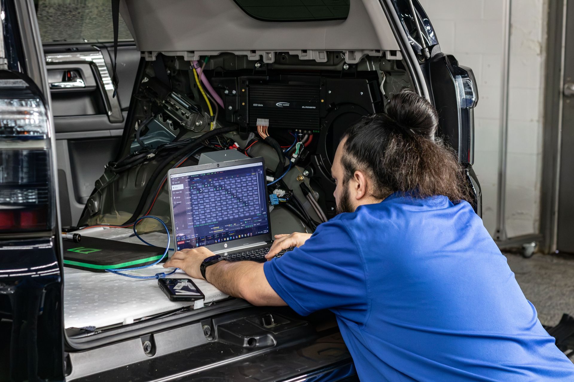 Technician in blue shirt using a laptop to diagnose wiring inside a vehicle service bay