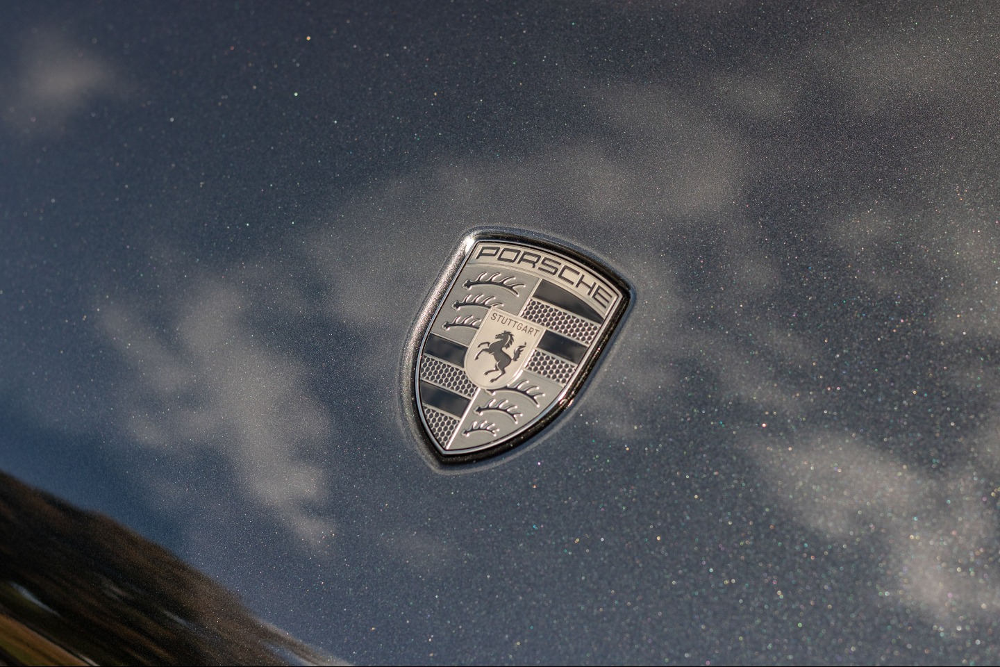 Porsche emblem on a metallic blue car hood, reflecting sky and light.