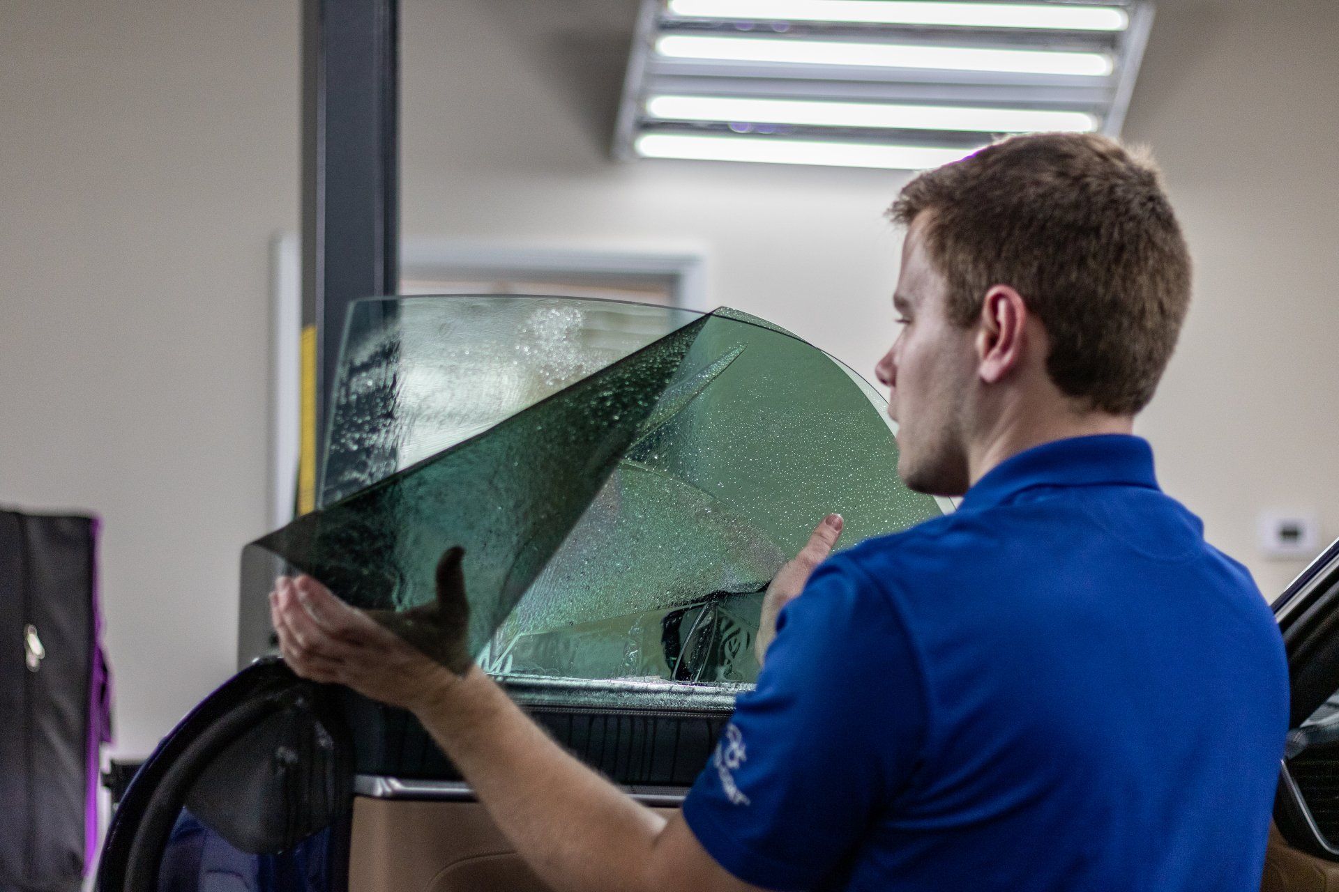 A man in a blue shirt is applying tinted glass to a car window.