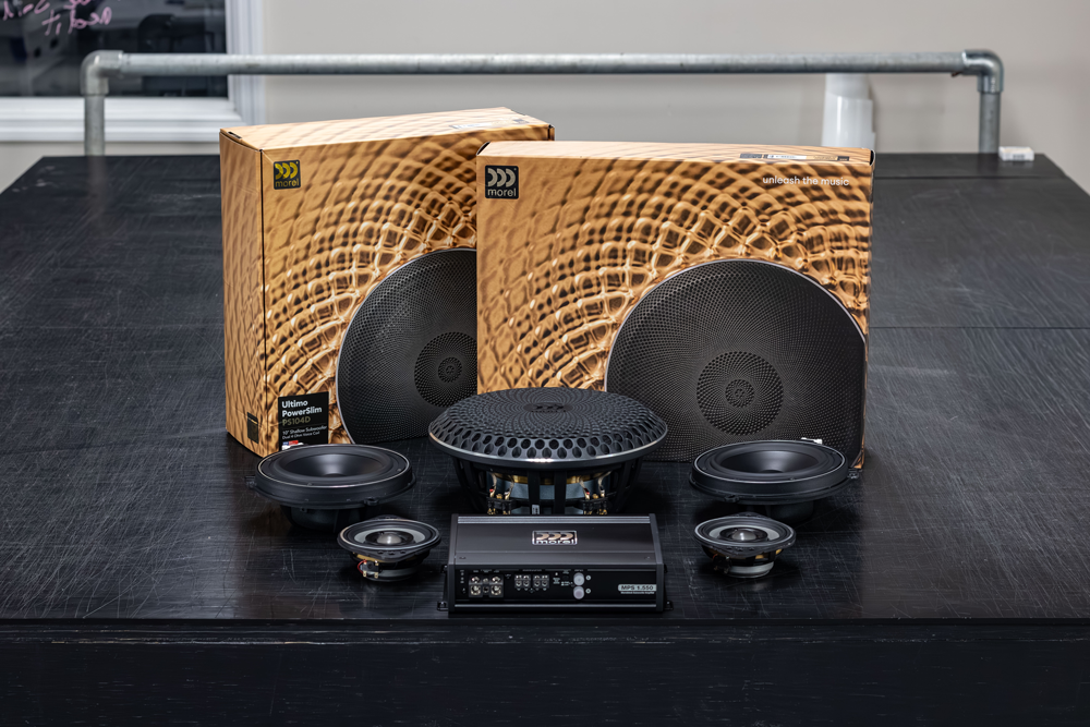 Car audio speaker components and subwoofer boxes arranged on a black table with wicker-backed cabinets.