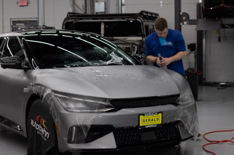A technician in a blue uniform applies protective clear film to the hood of a silver car in a bright workshop.