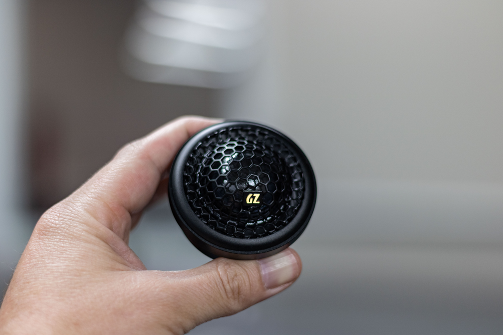 Hand holding a small black round speaker with a honeycomb grille and yellow logo on a blurred indoor background