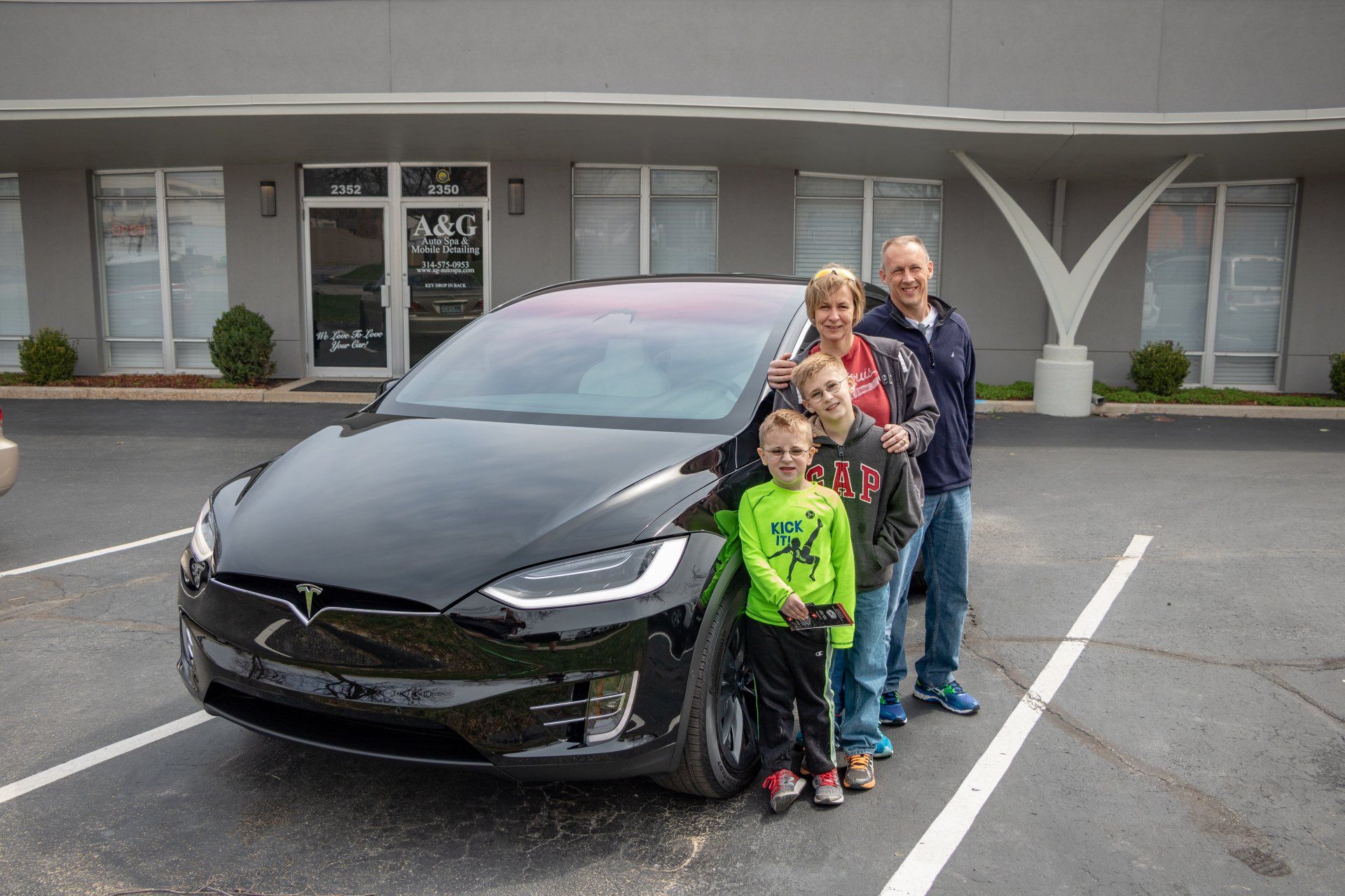 A family is standing in front of a black tesla model x in a parking lot.