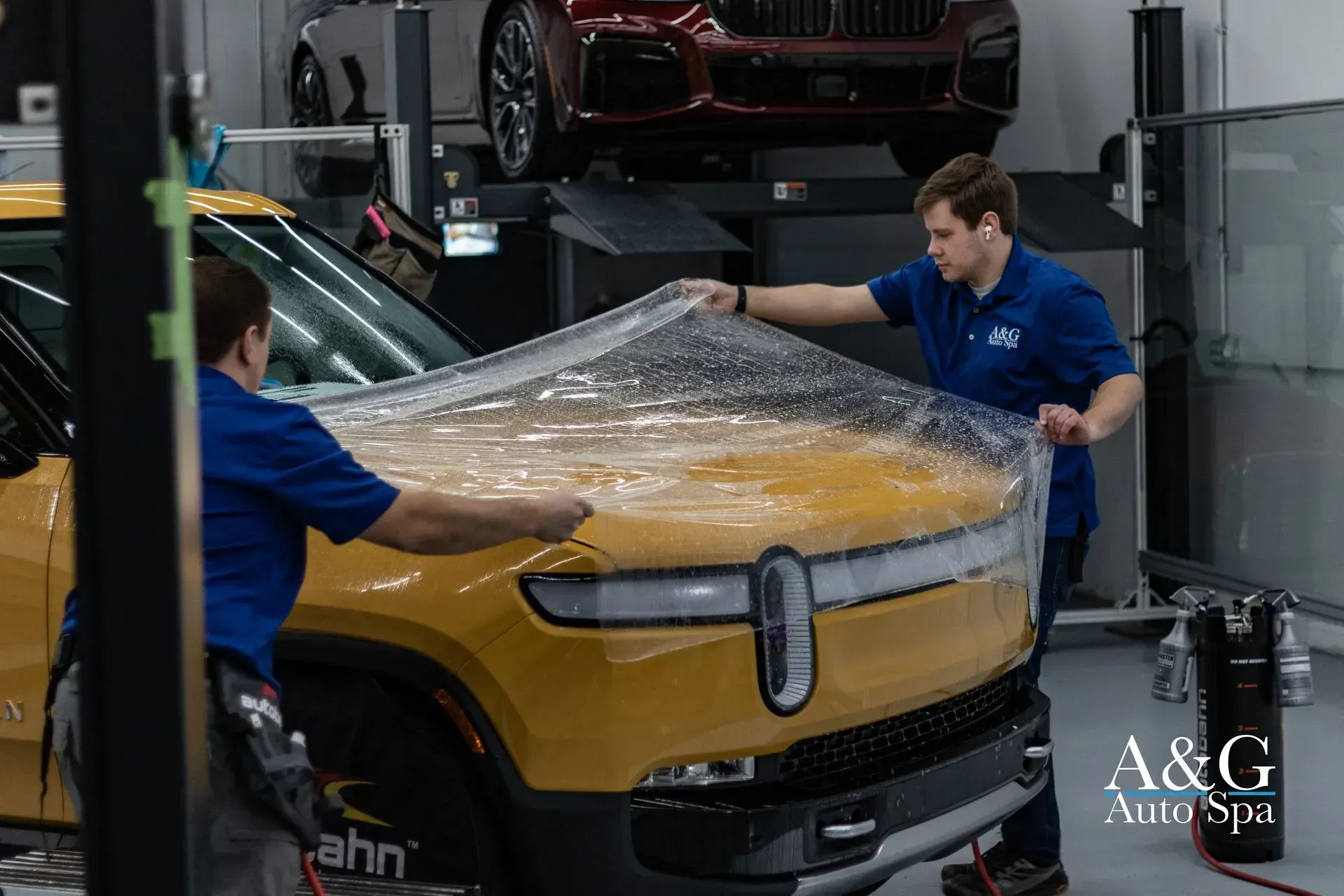 Two men are wrapping a yellow car in plastic in a garage.