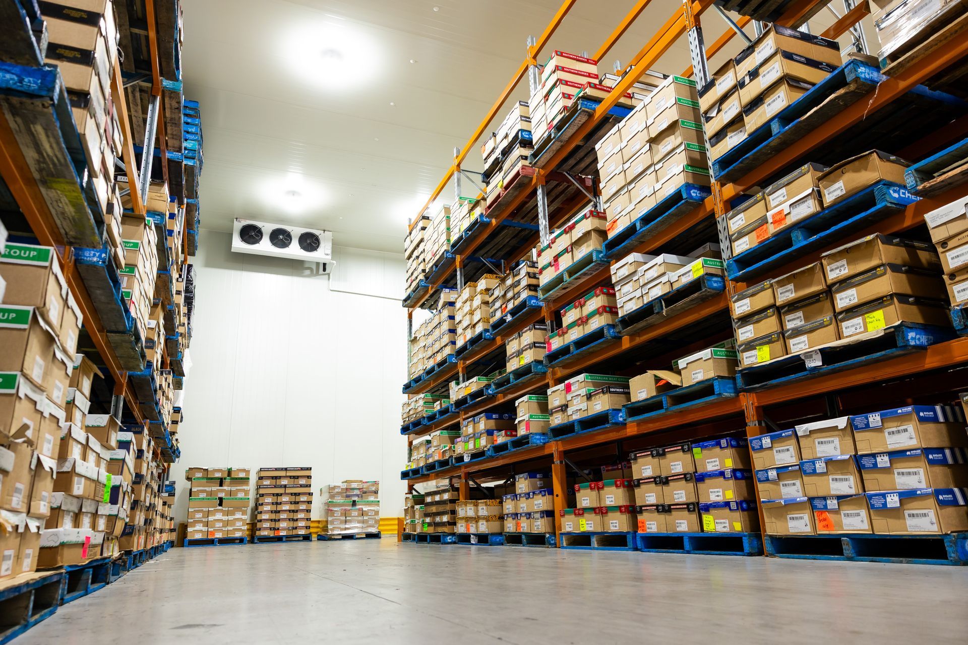Aisle view of a warehouse with tall shelves stacked with boxed inventory and a bright concrete floor.