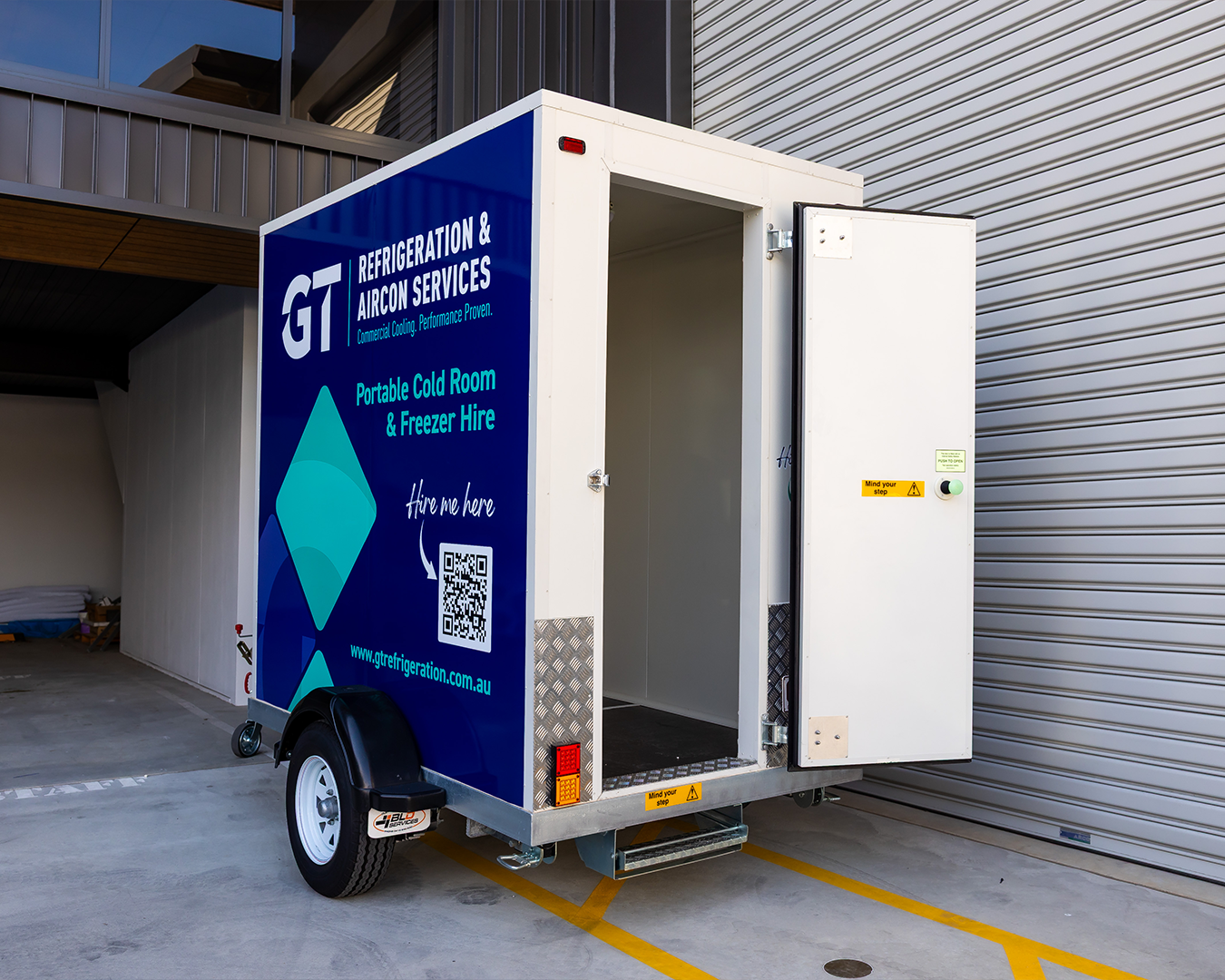 White utility trailer with blue branding parked by a loading dock, rear door open.