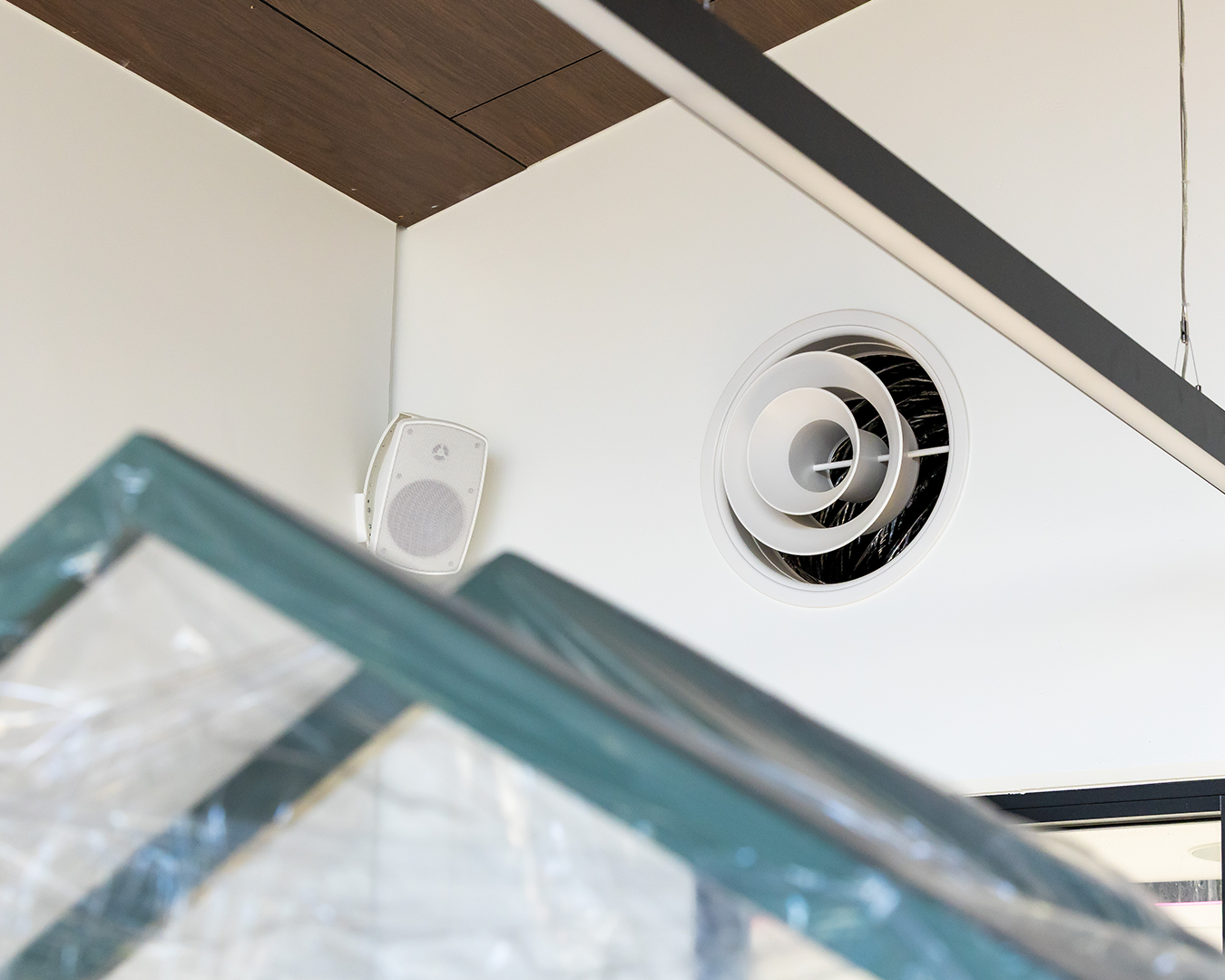 Ceiling corner with a round vent and small white speaker, partially obscured by a glass structure