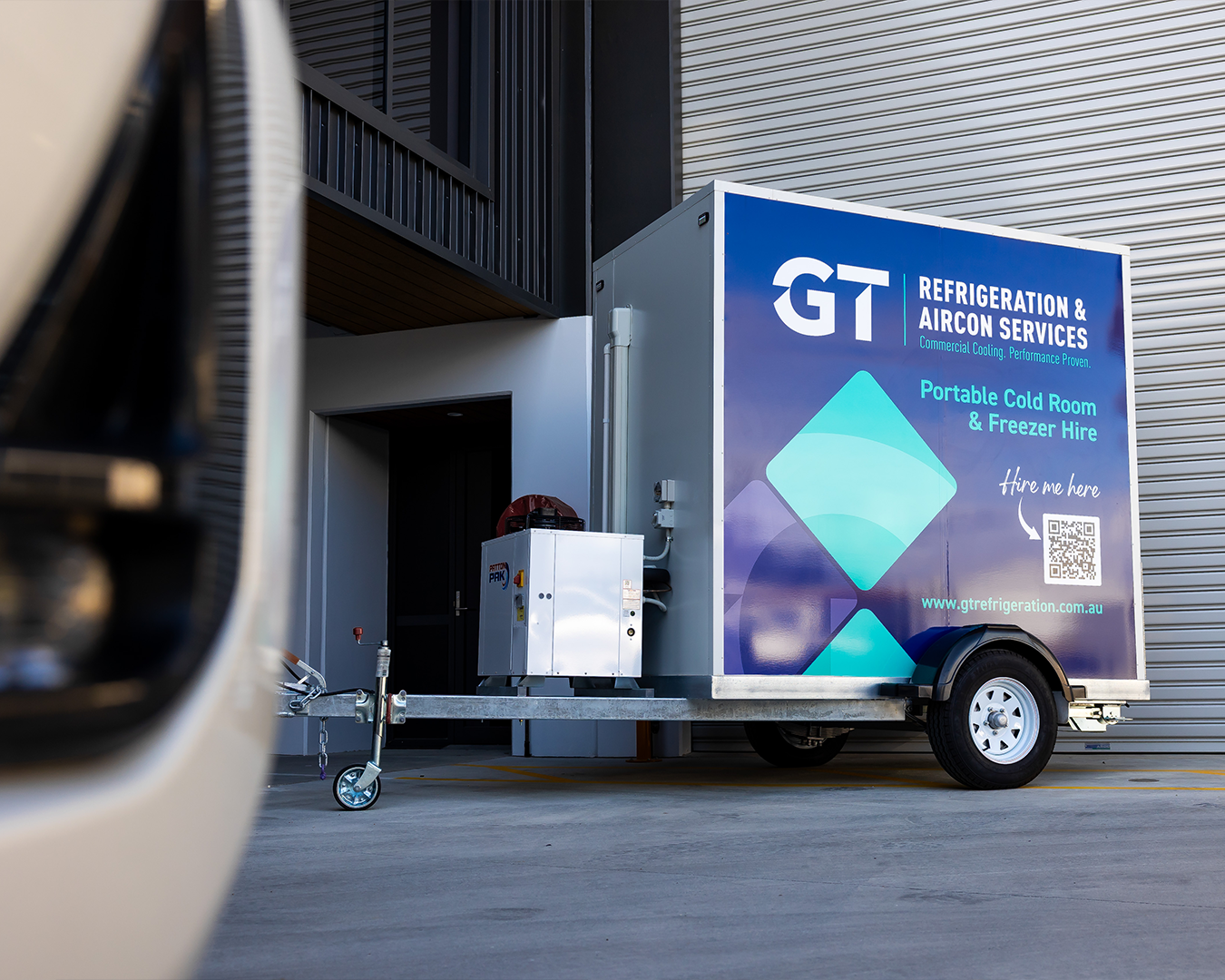 Small GT-branded service trailer parked beside a building, with a blue sign and wheel visible.