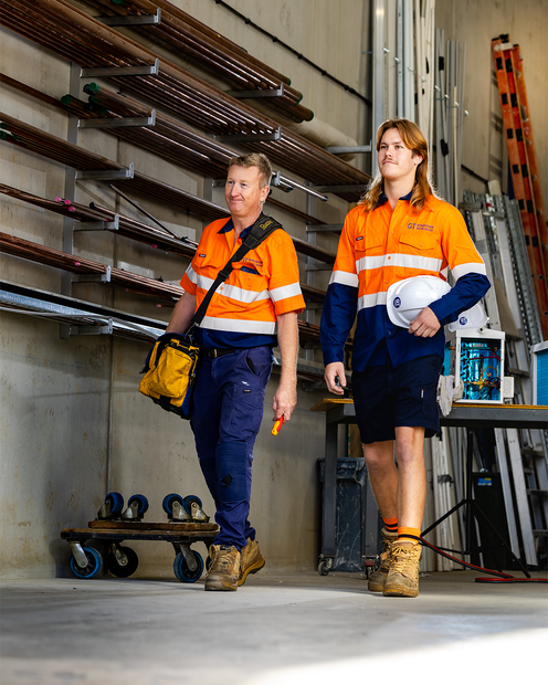 Two workers in orange safety shirts and blue pants walk inside a warehouse carrying hard hats.