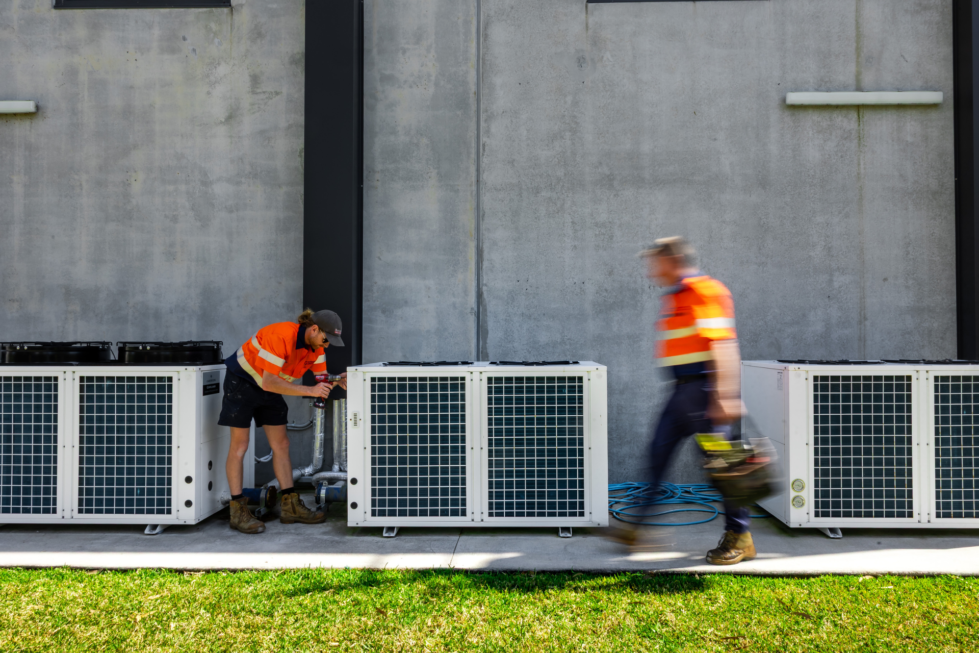 Workers in orange safety vests inspecting outdoor HVAC units against a concrete wall