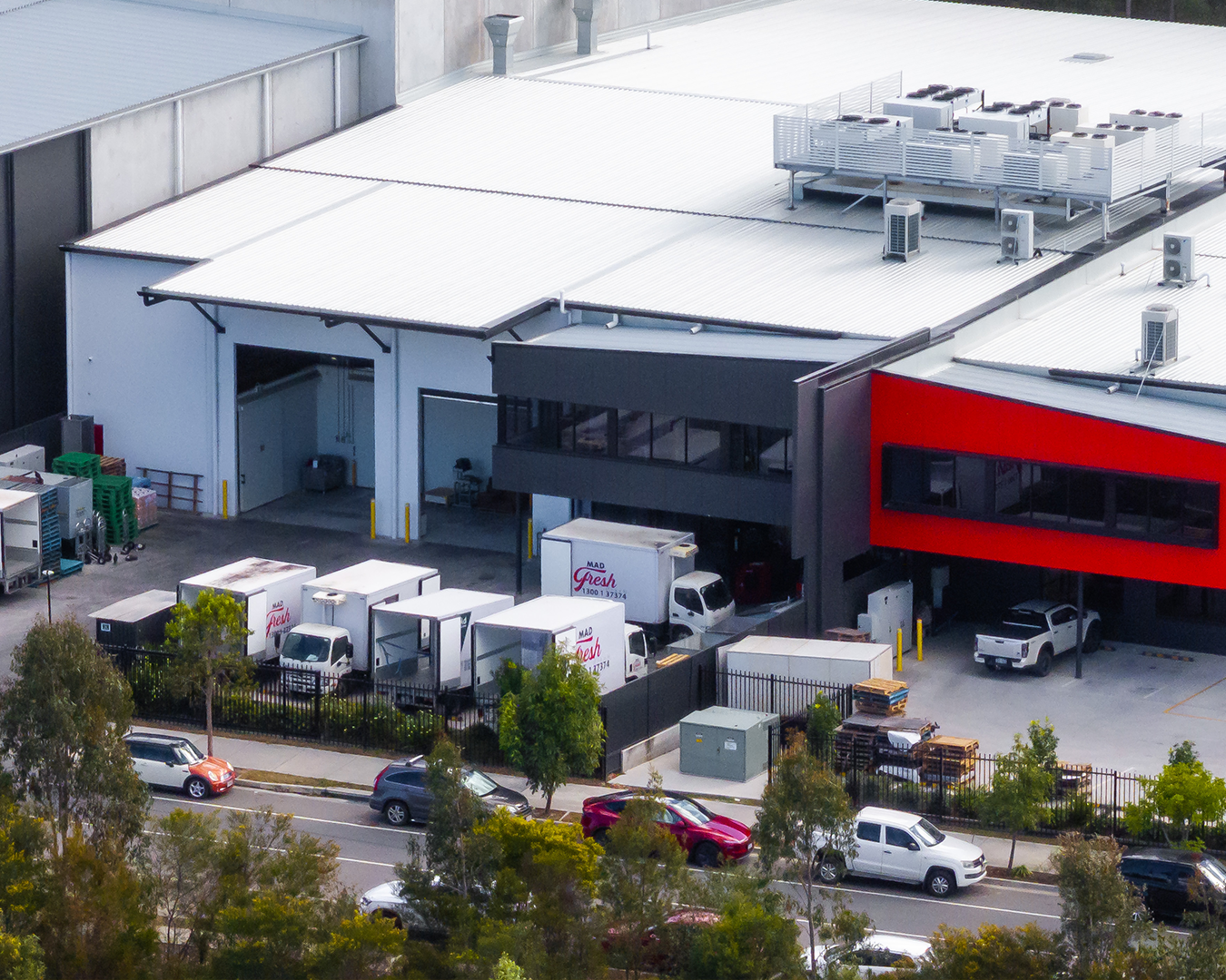 Industrial building with red-accented facade, parked trucks, and a road lined with trees.