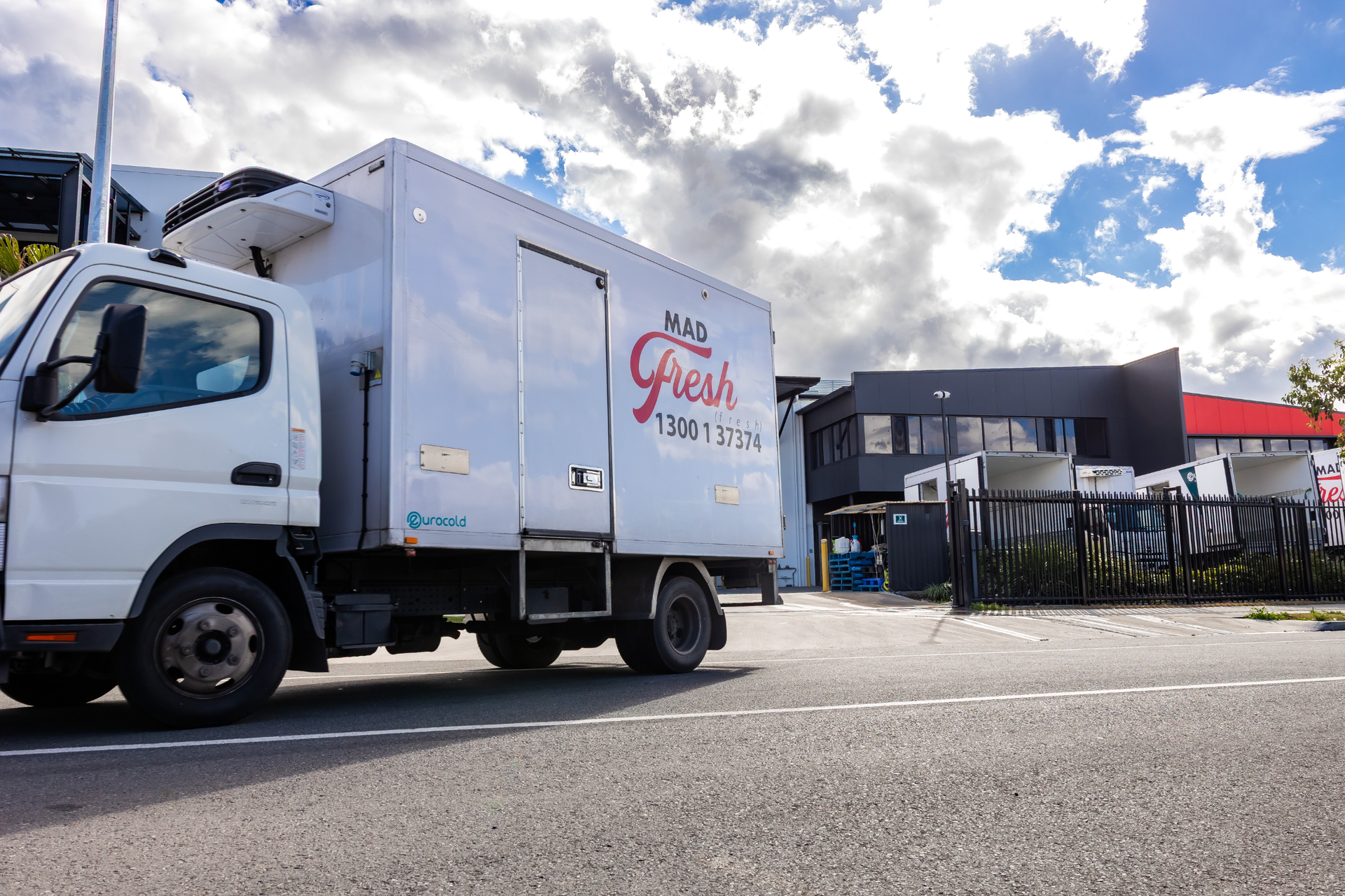 White delivery truck parked in a lot under a cloudy sky, with a building and fence in the background