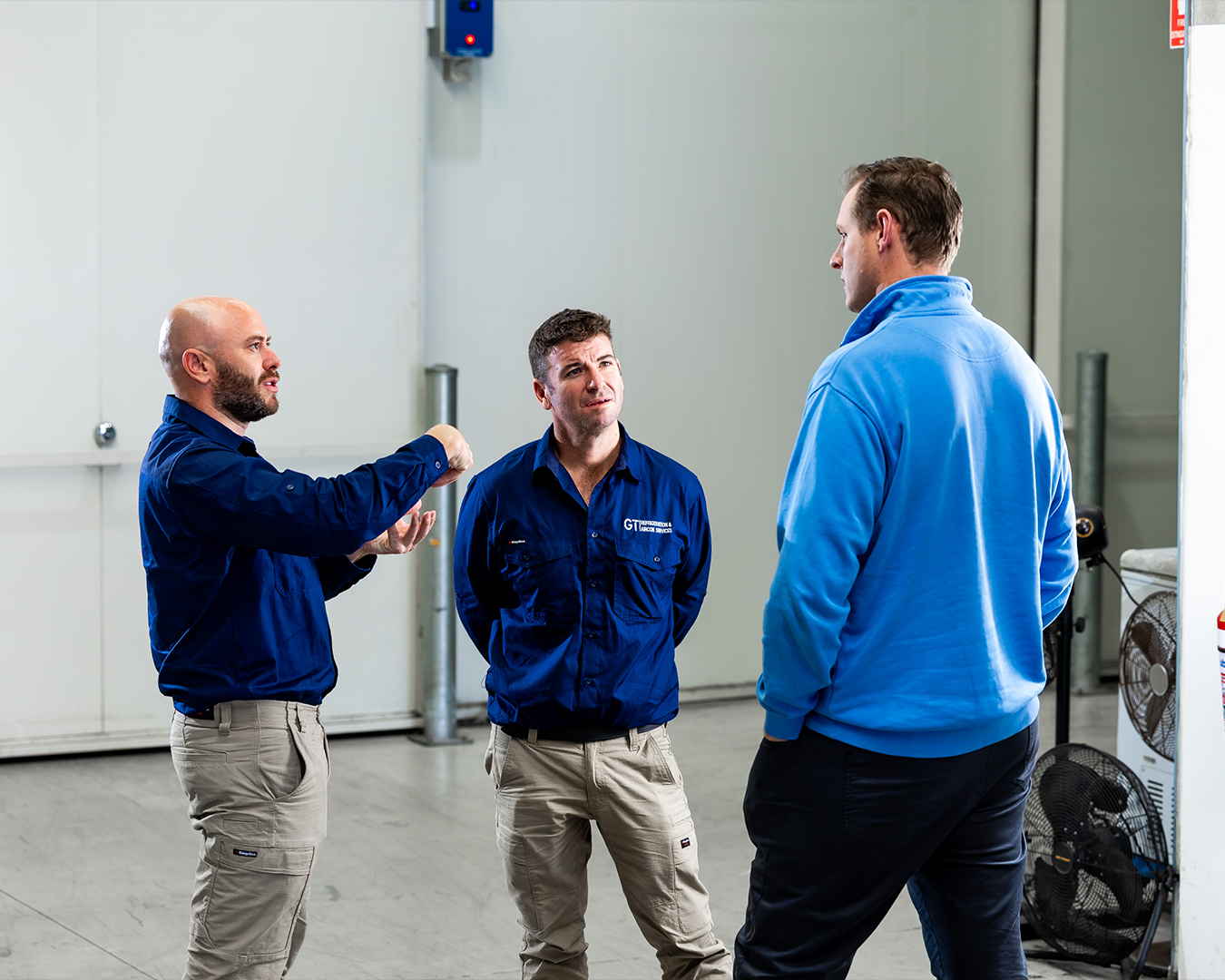 Three men in blue work jackets talk in an industrial warehouse.