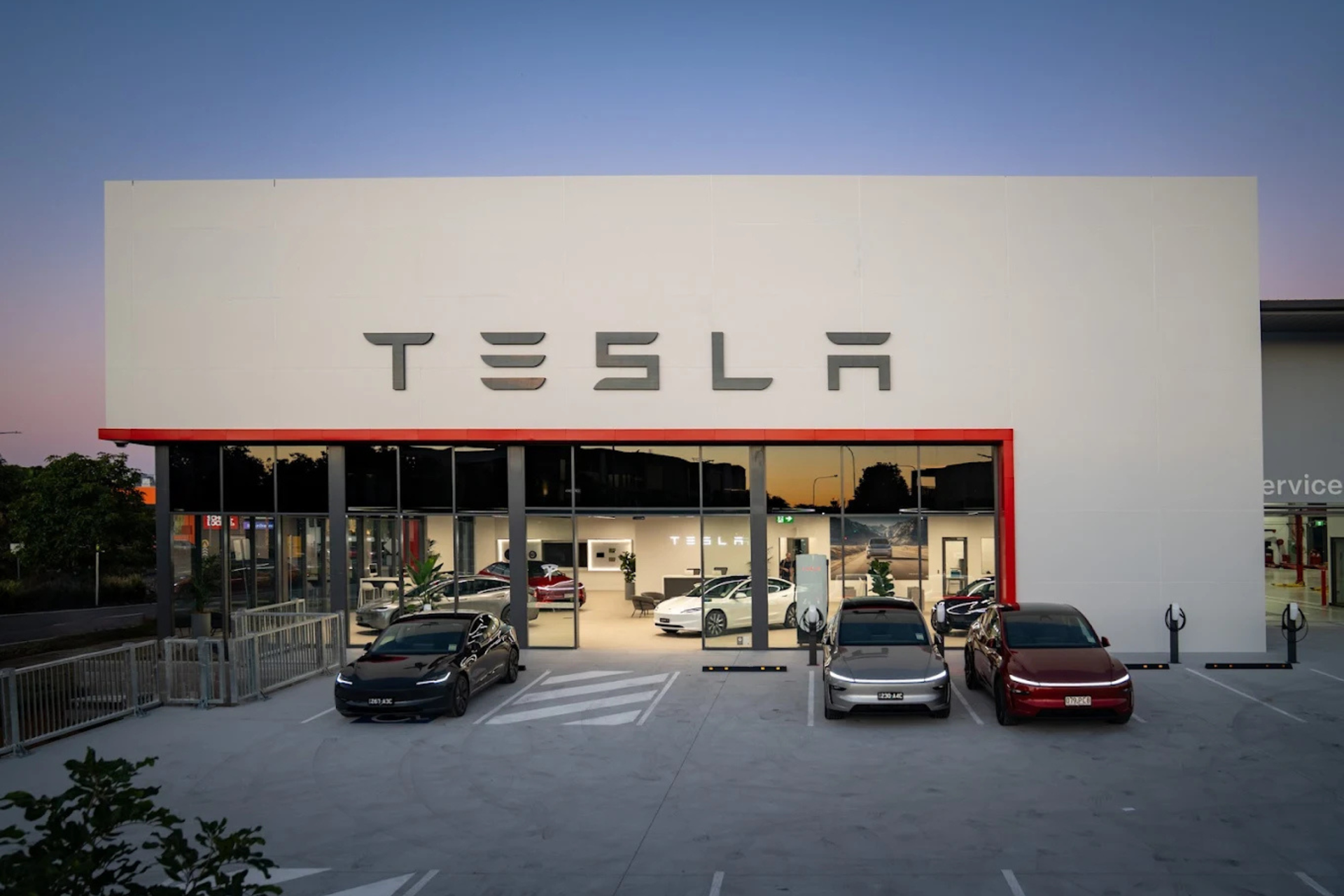 Tesla showroom exterior at dusk with three parked cars under a large white facade.