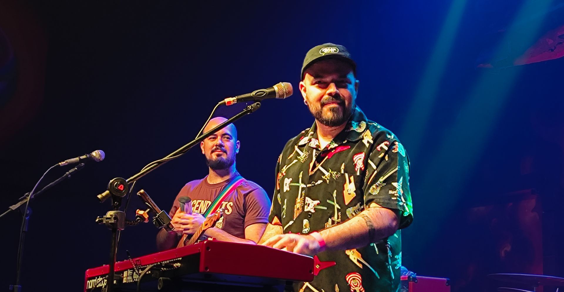 Integrante da banda Papangu toca teclado no palco enquanto o vocalista canta ao microfone.