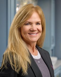 Smiling woman in a dark blazer standing in an office hallway, with long blonde hair.