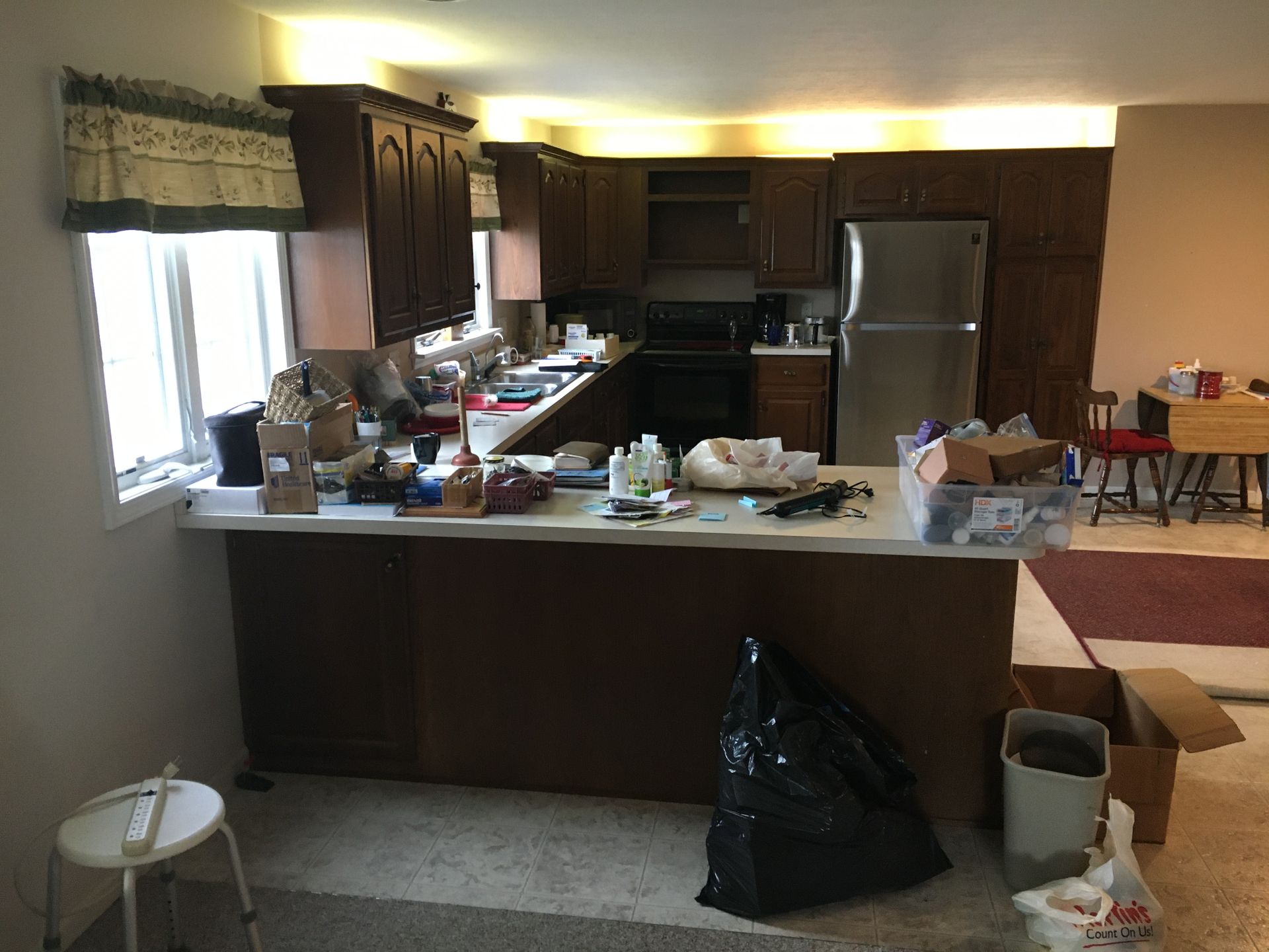 Cluttered kitchen with counters covered in items, cabinets, fridge, and a trash bag on the floor.