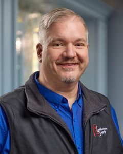 Smiling man in a blue shirt and dark vest, standing in a hallway.