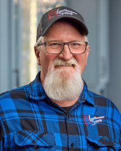 Man in blue plaid shirt and black cap, smiling in front of a blurred outdoor background