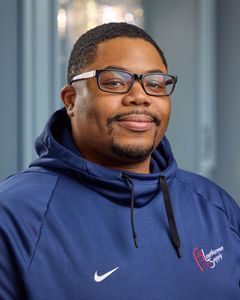 Smiling man in glasses and a blue hoodie against a blurred indoor background