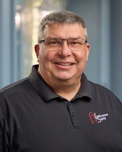 Smiling man in a dark polo shirt with glasses, standing in an office setting