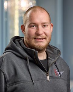 Smiling person in a gray hoodie, seated indoors against a softly blurred background.
