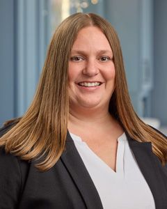 Smiling woman in a black blazer and white blouse standing indoors with a blurred office background