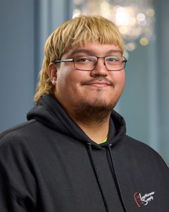 Smiling person with blond hair and glasses wearing a black hoodie against a blurred indoor background