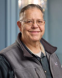 Smiling person in glasses and a gray jacket standing indoors against a blurred blue background