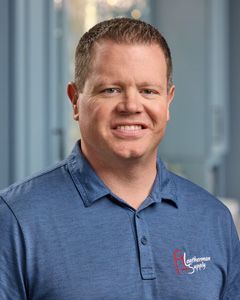 Smiling man in a blue polo shirt with a company logo, standing indoors against a blurred background