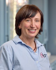 Smiling woman in a light blue collared shirt, standing in a blurred office hallway.