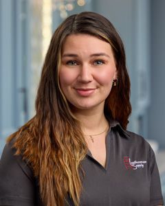 Portrait of a smiling woman with long brown hair in a dark gray polo shirt, standing outdoors near a modern building.