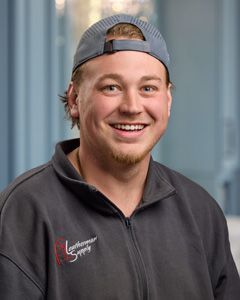 Smiling person in a gray cap and dark zip-up jacket against a blurred indoor background