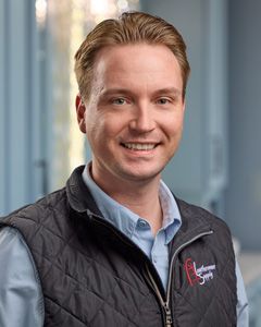 Smiling man in a black vest and blue shirt standing in a bright office hallway