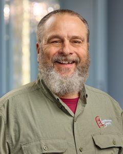 Smiling man with gray beard in olive shirt, standing in front of a blurred office background