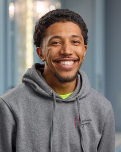 Smiling person in a gray hoodie standing indoors near a window