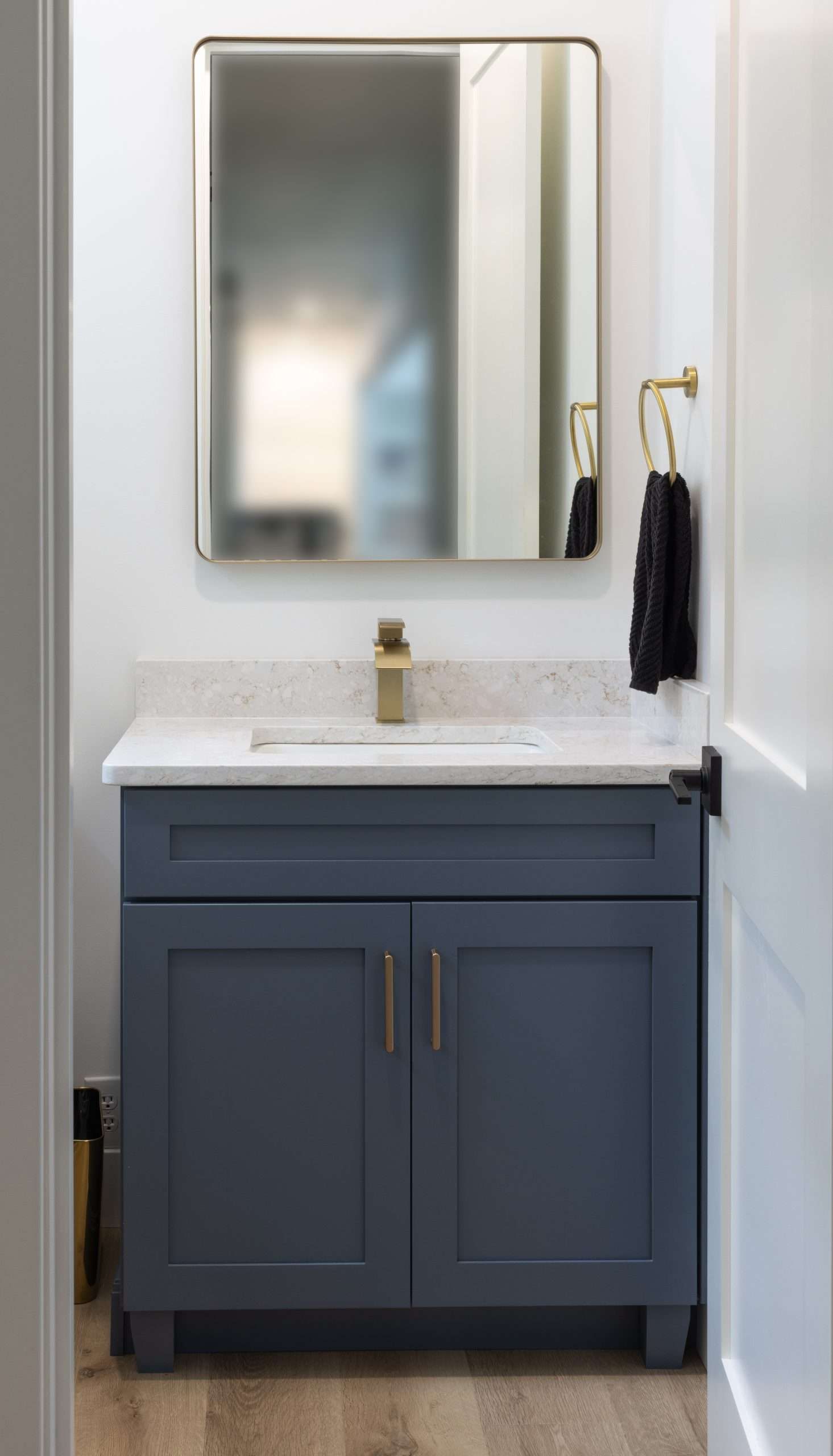 Blue bathroom vanity with white countertop, mirror, and gold faucet in a narrow white room