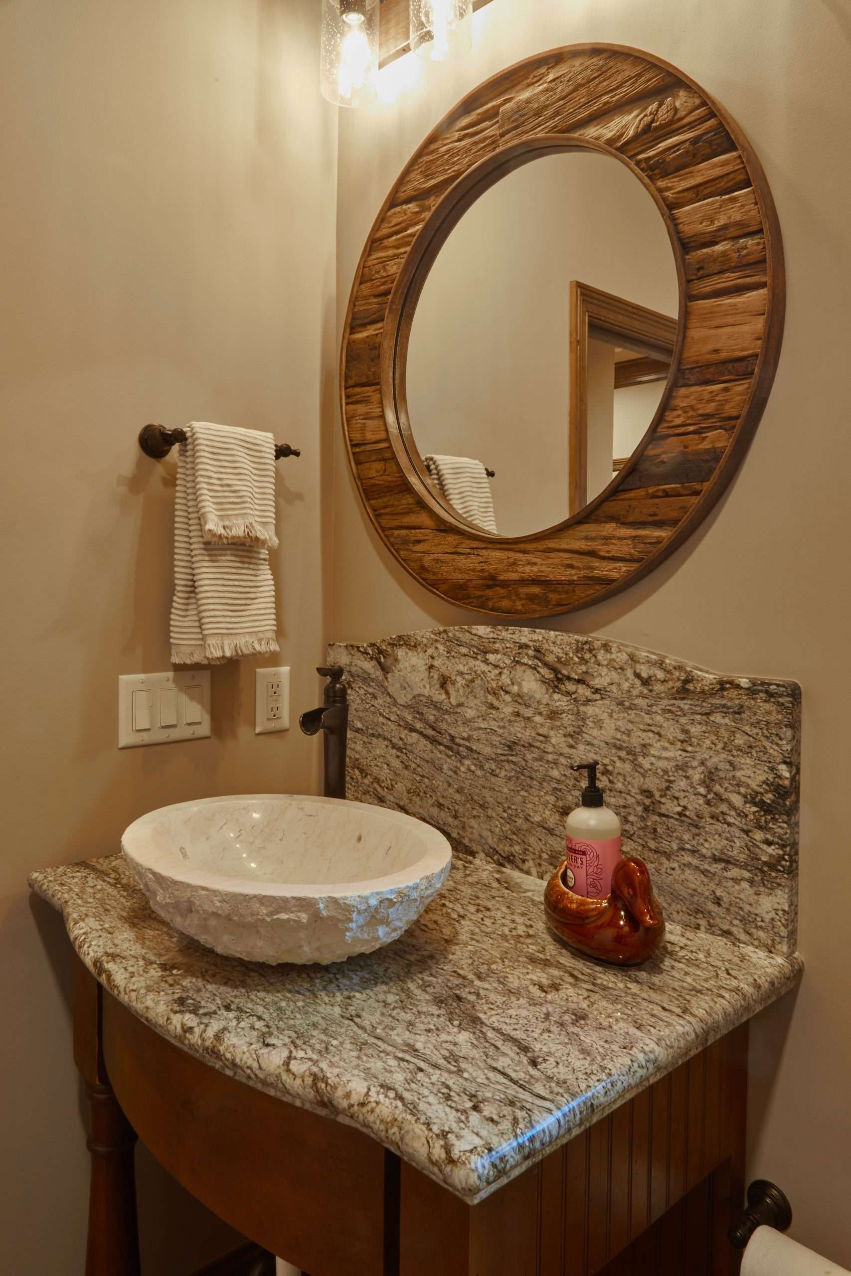 Small bathroom vanity with stone countertop, round woven mirror, and white vessel sink on a wooden cabinet.
