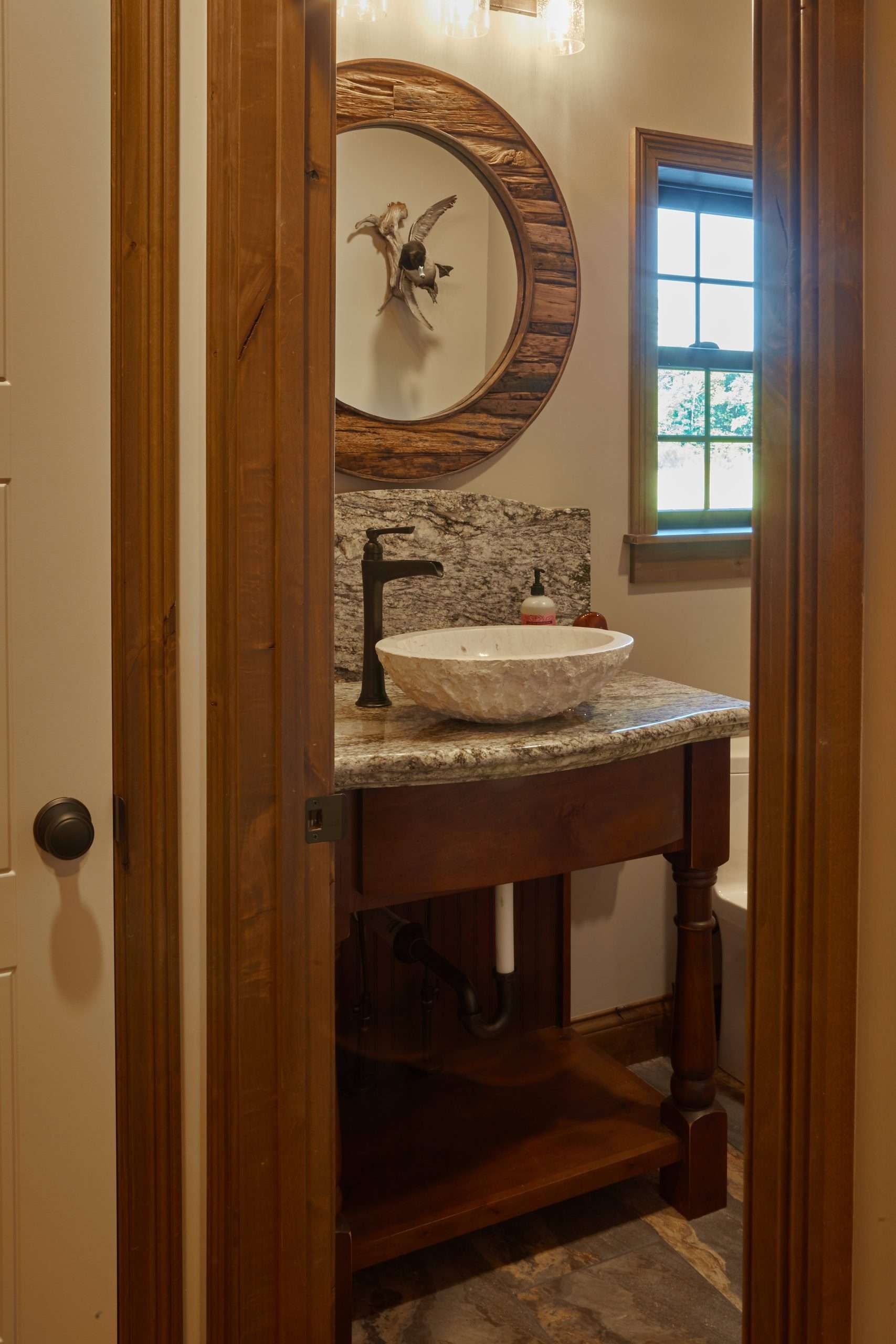 Small bathroom vanity with stone sink, round mirror, and window beside wooden doorframe