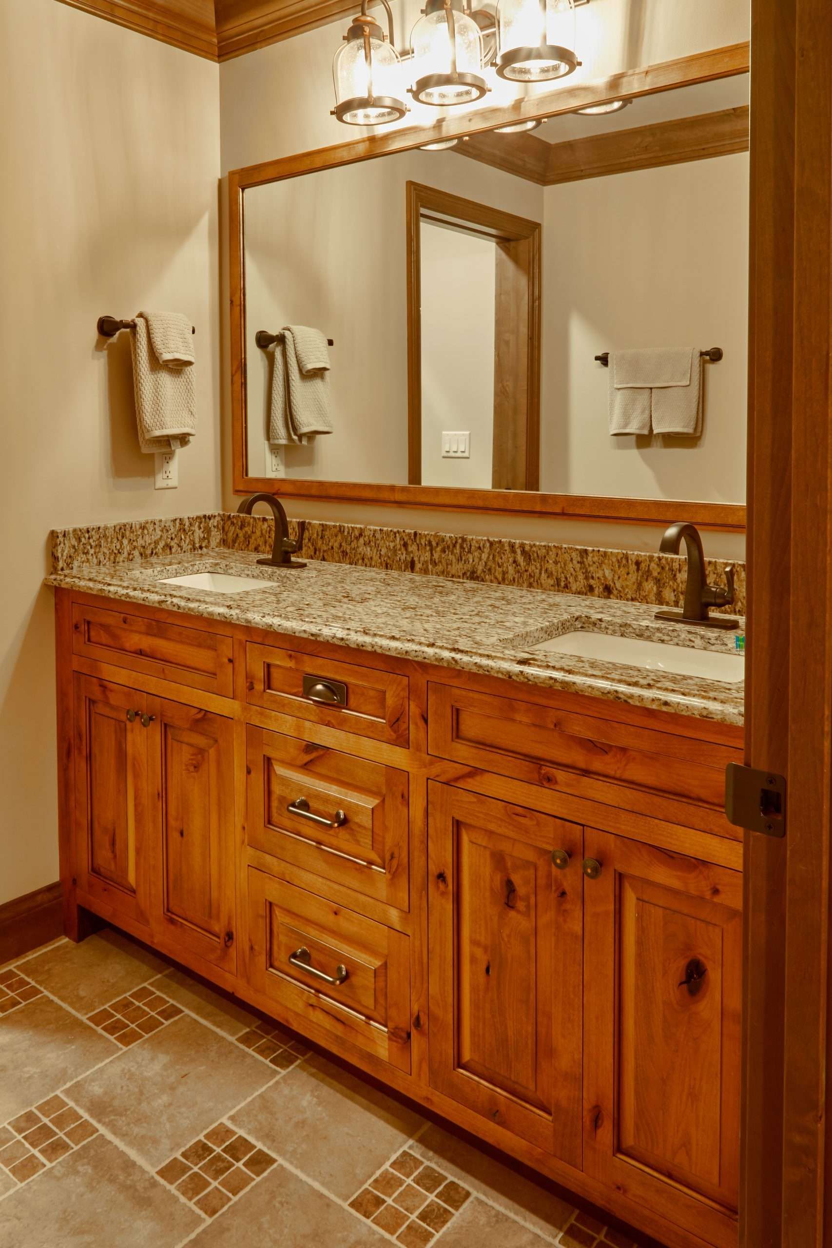 Wooden bathroom vanity with granite countertop, large mirror, and wall sconces in a tiled bathroom.