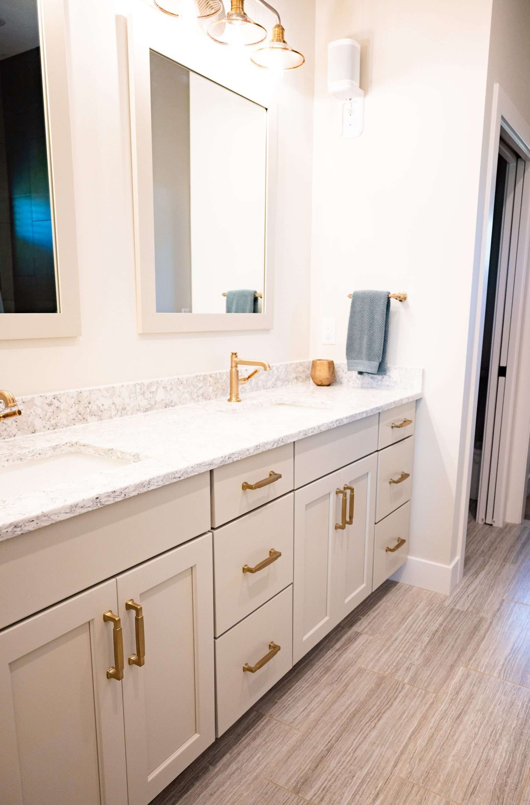 Bright bathroom vanity with white cabinets, gold fixtures, mirrors, and a tiled floor