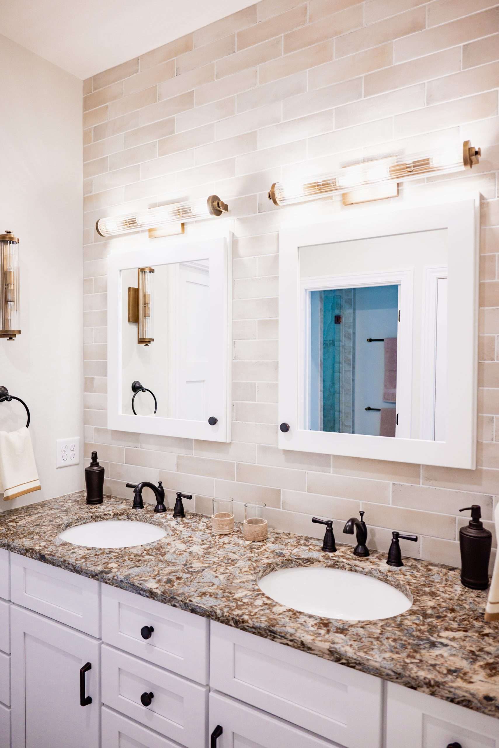 Bright bathroom vanity with granite counter, twin mirrors, and brass light fixtures against white tile walls