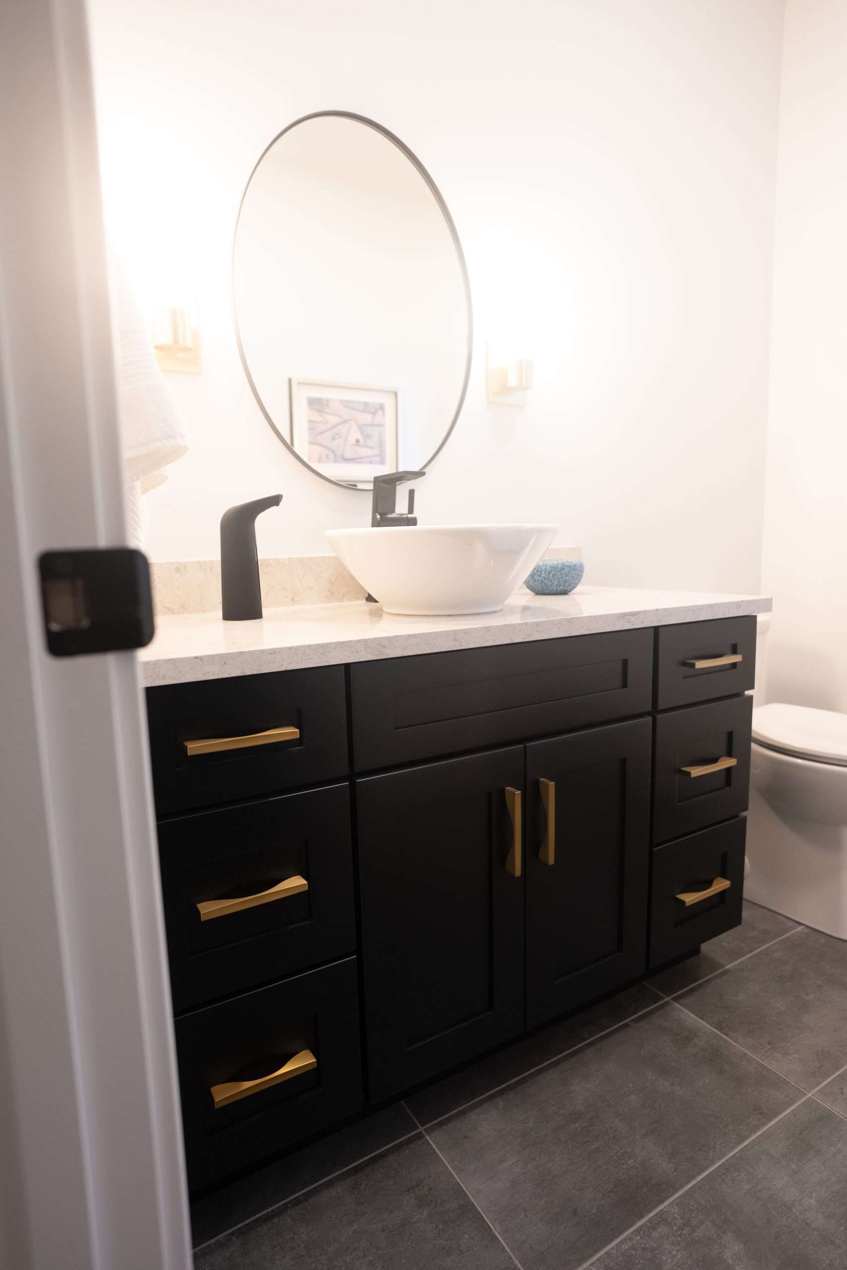 Black bathroom vanity with gold handles, white countertop, oval mirror, and dark tile floor.